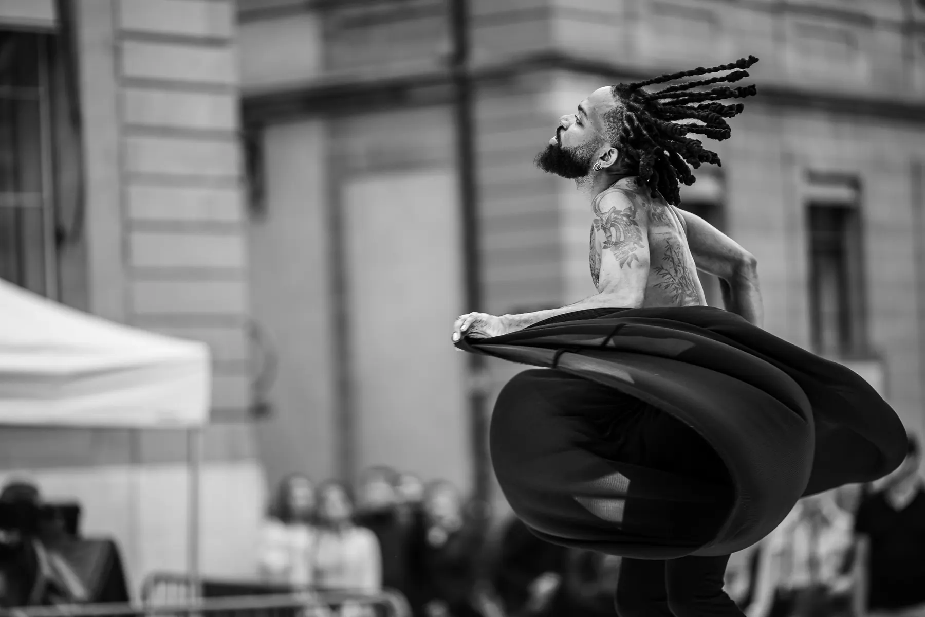 Ivan Larson en plein tour lors d'une performance en extérieur, un long tissu noir tournoyant autour de lui, photo en noir et blanc